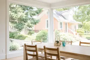 Wooden dining table and chairs beside floor-to-ceiling corner windows with a clear view of a house and garden outside